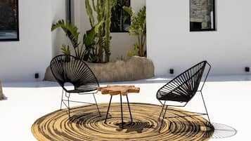 Una terraza en la azotea con dos sillas de estructura metálica negra, una mesa de madera y un tapete tejido. Se observan plantas en macetas y cuadros en las paredes.