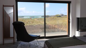 Ein Schlafzimmer mit einem großen Schiebeglasfenster, das Blick auf einen Strand bietet, einem schwarzen Sessel und einem Bett mit grüner Decke.