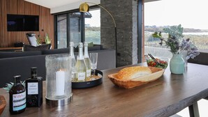 Ein modernes Wohnzimmer mit einer Holzwand, einem großen Fenster und Blick ins Freie. Es gibt ein Sofa, einen Couchtisch mit einer Weinflasche, Gläsern und einer Vase mit Blumen.