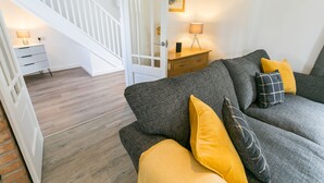 A living room with a grey sofa, yellow cushions, a wooden floor, a white staircase, and a small bedside table with a lamp.