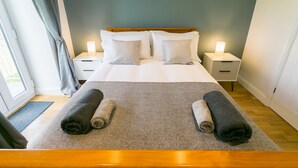 A neatly made bed with two pillows and two rolled towels, a bedside table with a lamp, and a window with curtains.