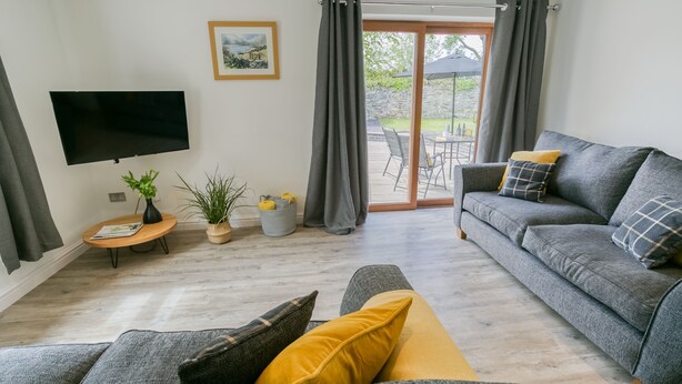 A living room with a grey sofa, a flat-screen TV, a small round table with a plant, and a sliding glass door leading to an outdoor patio.