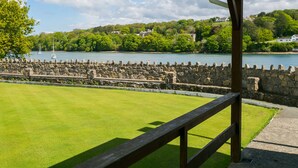 A grassy area with a stone wall and a wooden railing, overlooking a body of water and a hilly landscape with buildings. - 10 Beach Road -  a sea view that sleeps 4 guests  in 2 bedrooms (Menai Bridge)