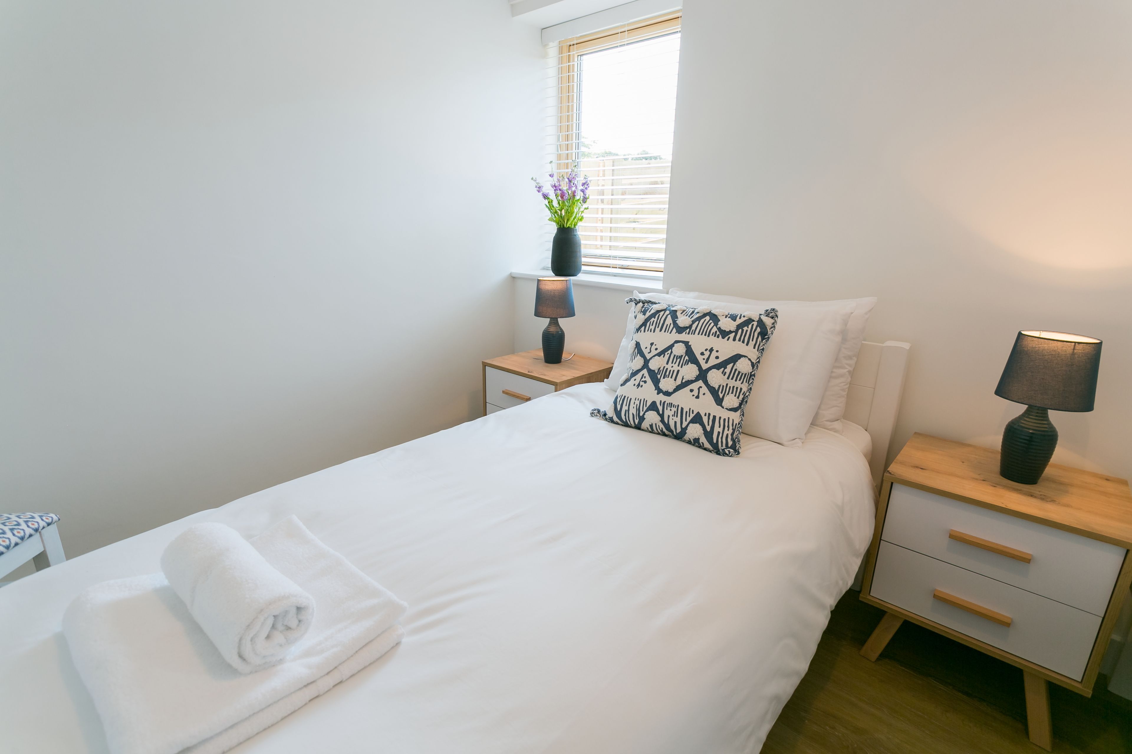 A neatly made bed with a white duvet, a patterned pillow, and a rolled white towel on the side. There are two bedside tables with lamps, one on each side of the bed.