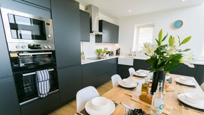 A modern kitchen with a built-in oven, microwave, and a dining area with a wooden table set for two. - Bwncath -  a coastal that sleeps 6 guests  in 3 bedrooms (Llanfairynghornwy)