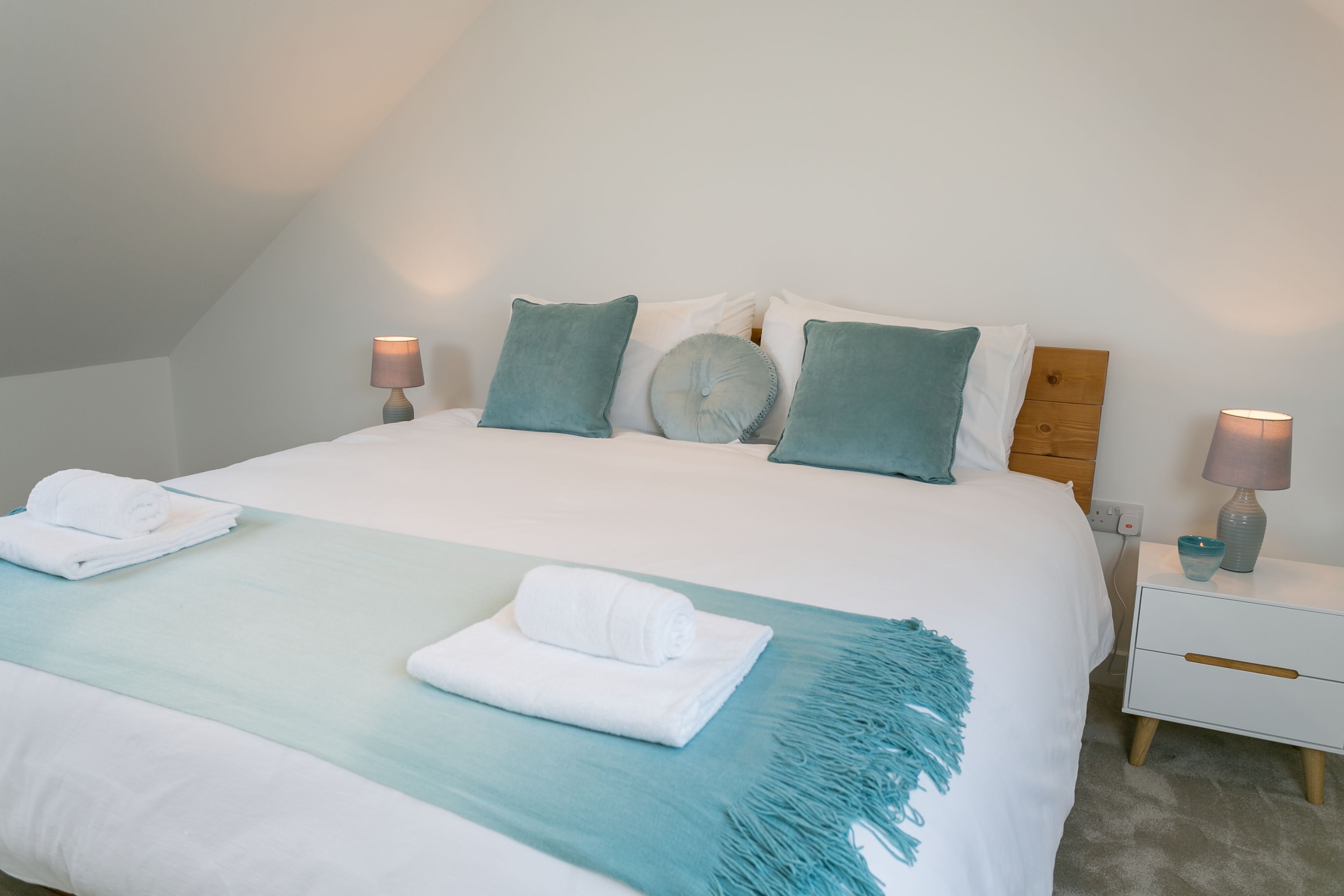 A neatly made bed with white linens and teal accents, including pillows and a throw blanket. There are two bedside tables with lamps and a small glass bowl on one.