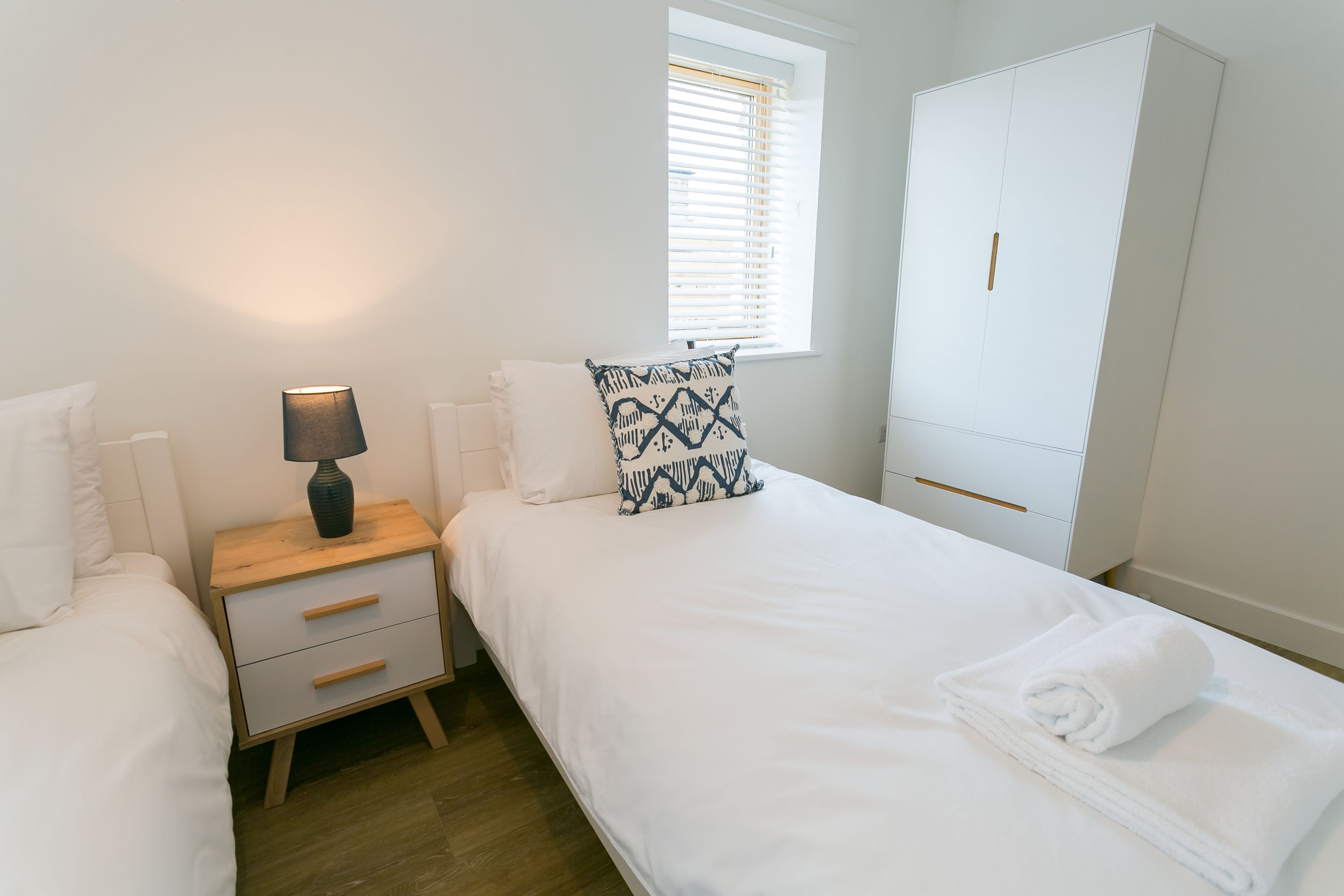 A bedroom with a bed, a nightstand, a lamp, a window with blinds, and a wardrobe.