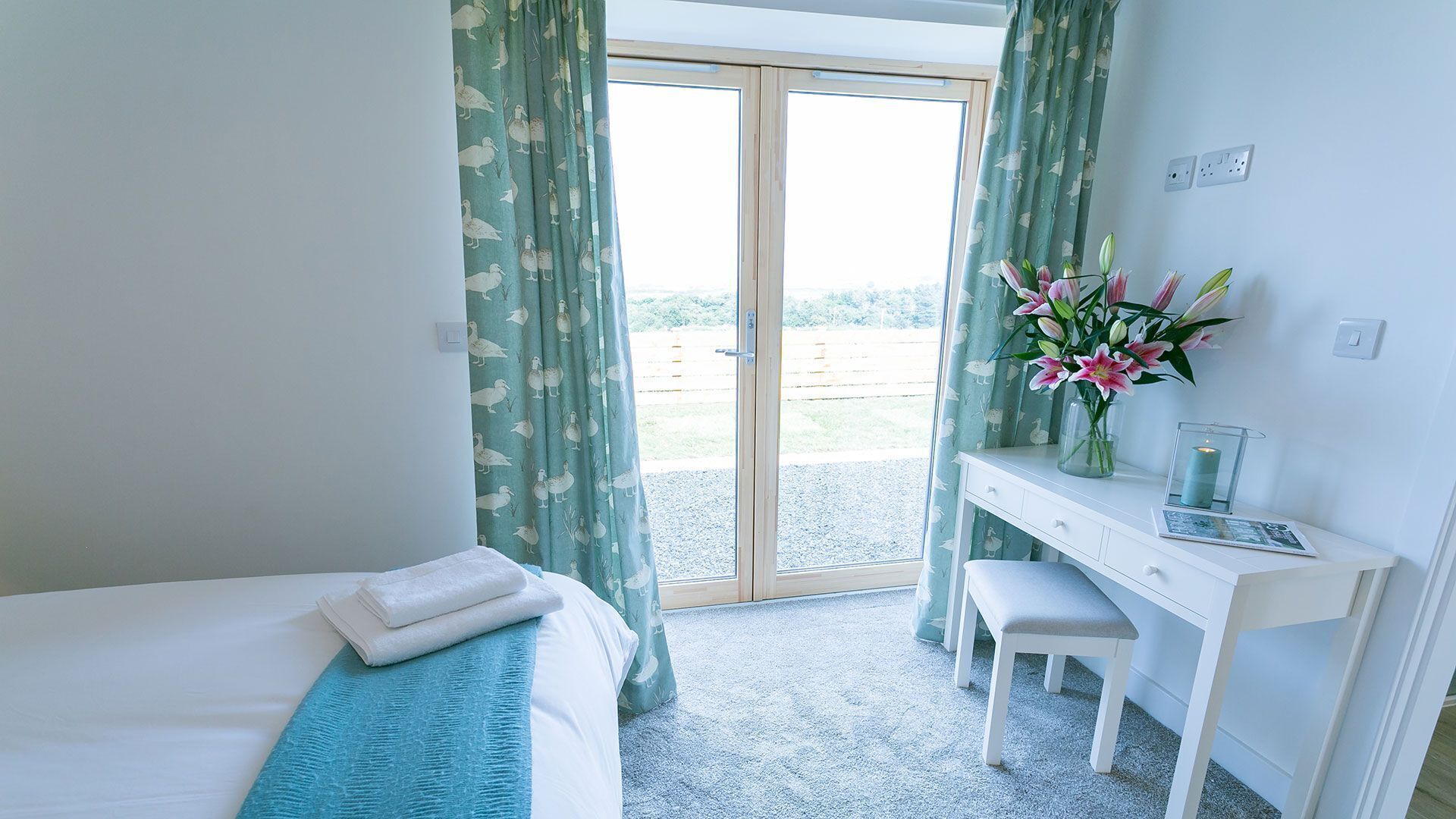 A bedroom with a bed, a desk, a chair, a vase with flowers, and a view of the outdoors.