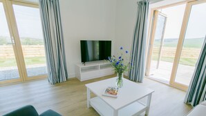 En moderne stue med et fladskærms-tv på et hvidt stativ, et sofabord med et magasin, en vase med blomster og en sofa.