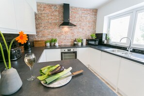 Una cocina moderna con armarios blancos, pared de ladrillo, campana extractora negra, ventana y encimera con verduras y una copa de vino.