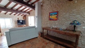 A living room with a stone wall, a wooden ceiling, a light blue sofa, a wooden console table, and a television. - Chambre Dans Maison de Caractère Proche du Mt Ventoux (Crillon-le-Brave)