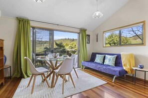 A modern living room with a glass dining table, white chairs, a purple sofa, and a view of a green landscape. - Kundaniwas - Lake Okareka Tiny House, Kayaks (Rotorua)