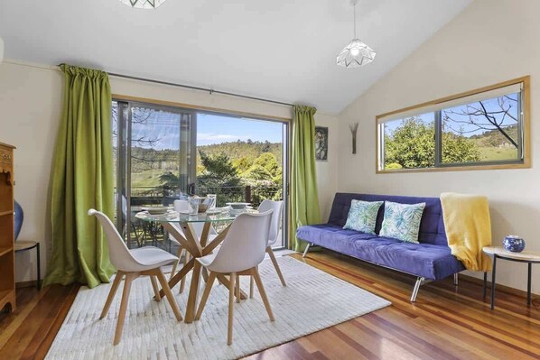 Un salon moderne avec une table à manger en verre, des chaises blanches, un canapé violet et une vue sur un paysage verdoyant.
