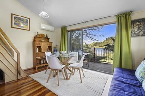 A dining area with a glass table and chairs, a wooden cabinet, and a view of a hillside through large windows. - Kundaniwas - Lake Okareka Tiny House, Kayaks (Rotorua)