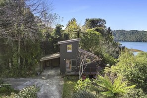 A small, single-story cabin with a sloped roof, surrounded by dense vegetation and a lake in the background. - Kundaniwas - Lake Okareka Tiny House, Kayaks (Rotorua)