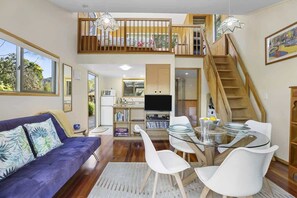 A modern living room with a sofa, a glass dining table, and a staircase leading to an upper level. - Kundaniwas - Lake Okareka Tiny House, Kayaks (Rotorua)