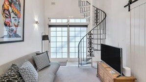 A modern living room with a grey sofa, patterned cushions, a flat-screen TV on a wooden stand, a black metal spiral staircase, and a large window with a view of the outside. - 'Latitude' - Stunning Opera House Views, Designer 2 Bedroom Executive Apartment w/ airconditioning (Kirribilli)