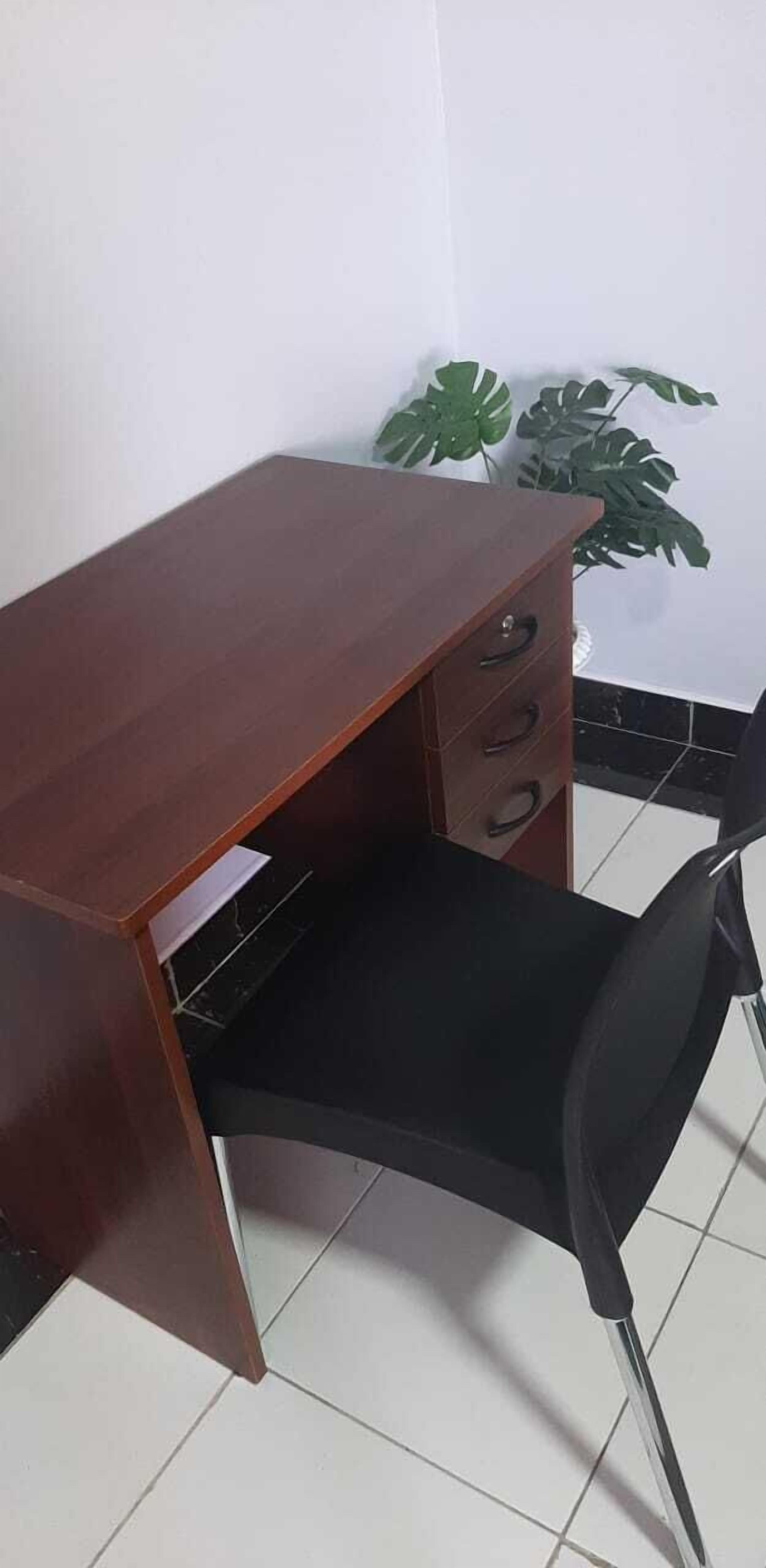 A wooden desk with drawers and a black chair in a room with tiled flooring and a potted plant.