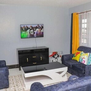 Una sala de estar con un televisor de pantalla plana montado en la pared, un mueble de entretenimiento negro, una mesa de centro blanca y dos sofás azules con cojines de colores.