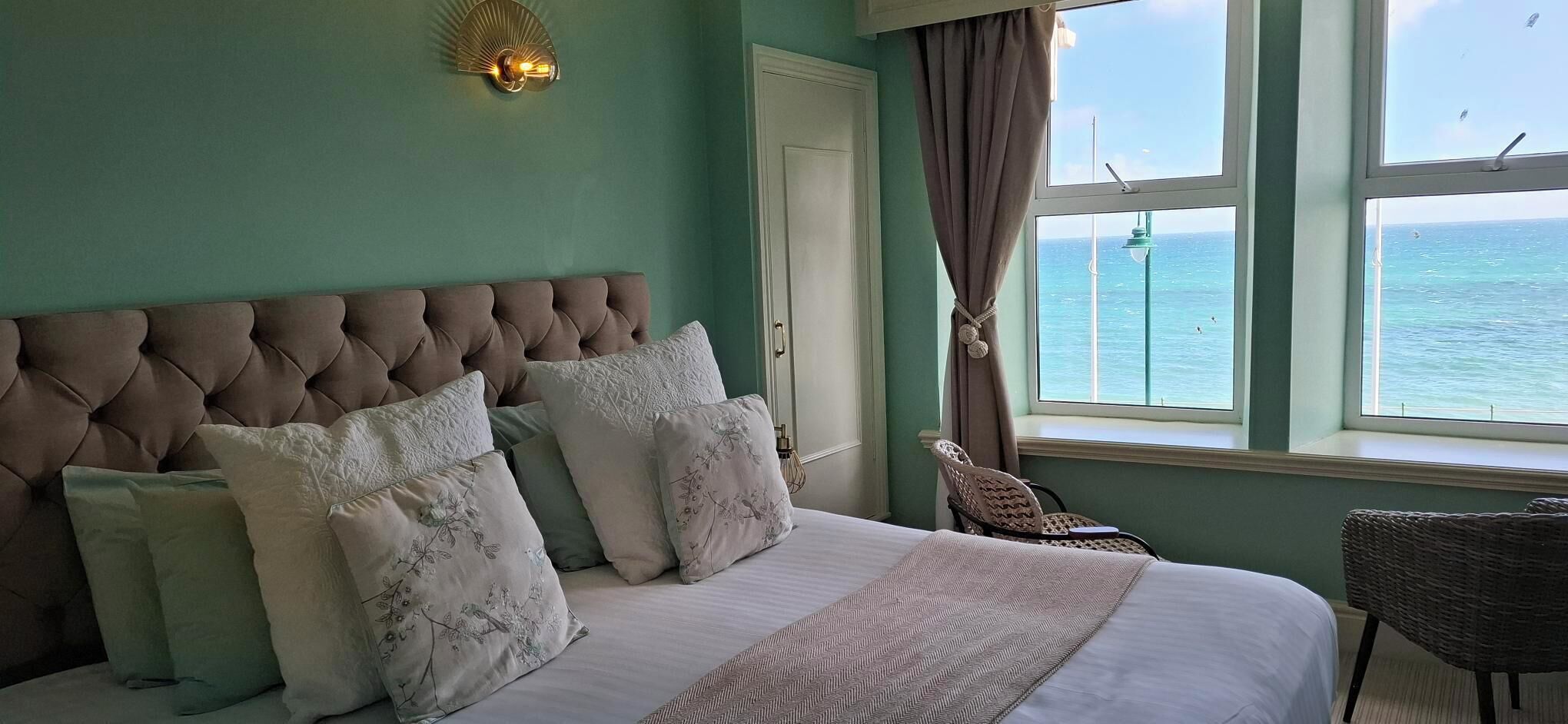 Ein Schlafzimmer mit einer gebogen Kopfstütze, einem Bett mit vielen Kissen, einem Fenster mit Meerblick und einem Korbsessel.
