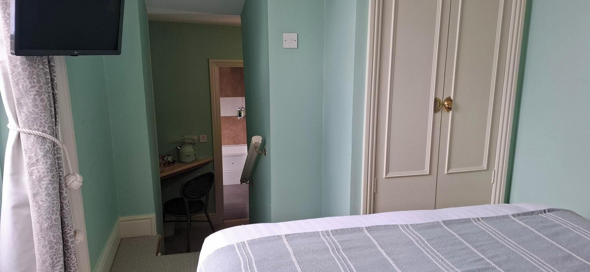 Un dormitorio con cama, televisor montado en la pared, escritorio y silla, y una puerta.