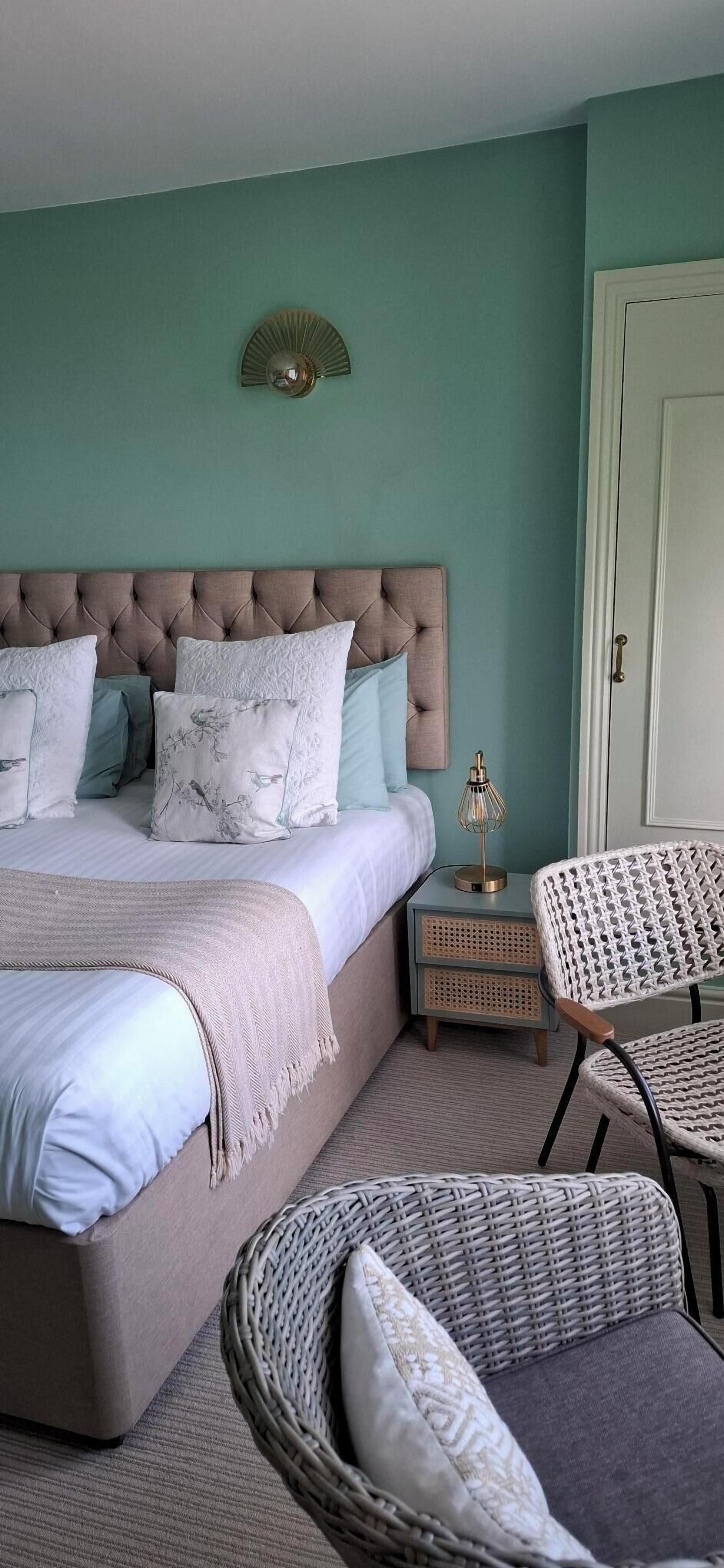 A bedroom with a tufted headboard, a bed with patterned pillows, a bedside lamp, wicker chairs, and a door.