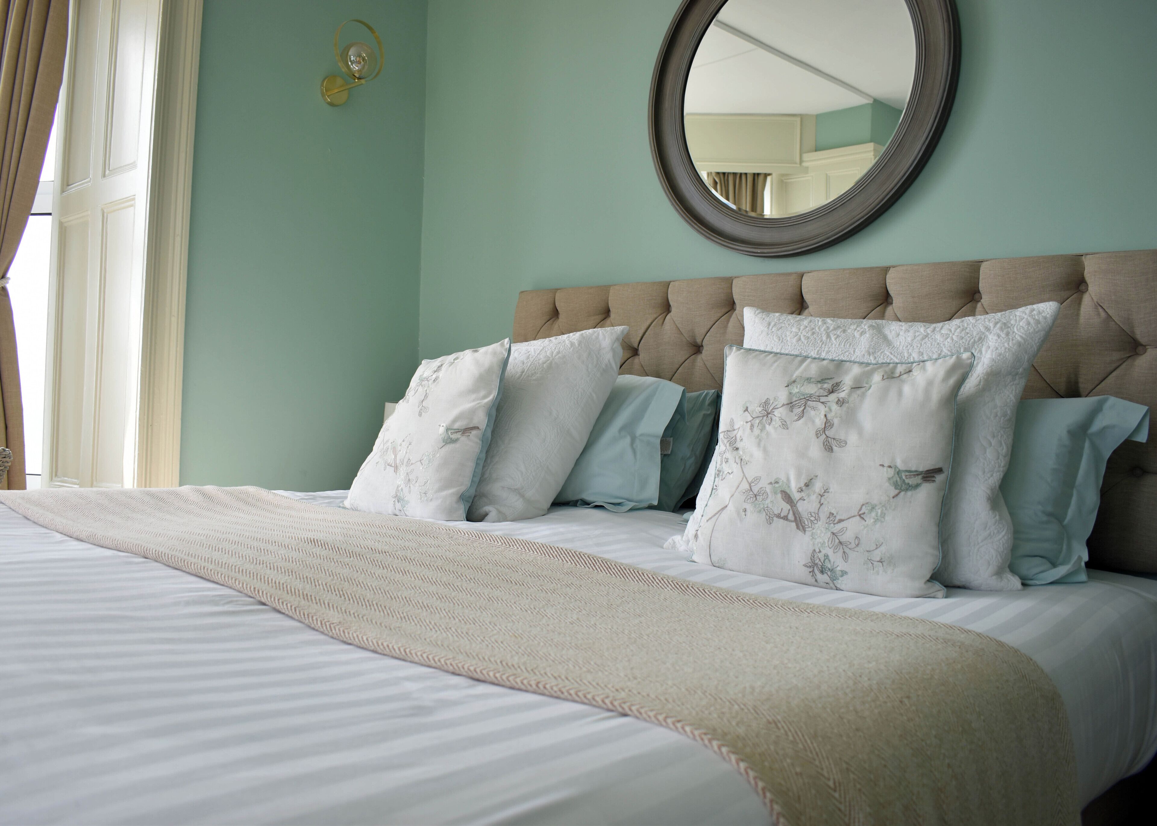 A neatly made bed with a tufted headboard, a round mirror above it, and a window with curtains on the left.