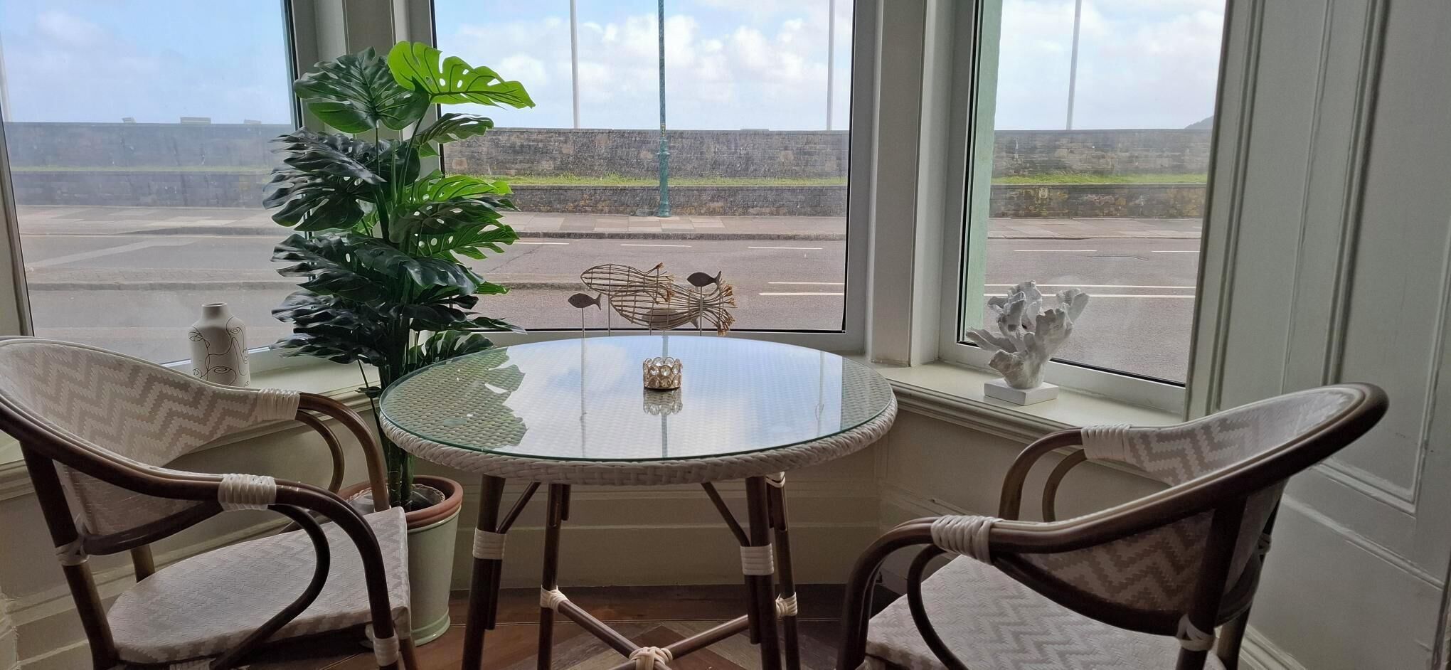 A room with a round glass table, two wicker chairs, a potted plant, and a view of a parking lot through large windows.