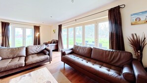 A spacious living room with two brown leather sofas, a wooden coffee table, and large windows offering a view of a green lawn and a building in the distance.