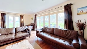 A spacious living room with two brown leather sofas, a wooden coffee table, and large windows offering a view of a green lawn and a building in the distance. - Stunning Coastal Cottage - Sleeping 8 in 4 Bedrooms - Pool and Beach Access Included. (Primrose Valley)
