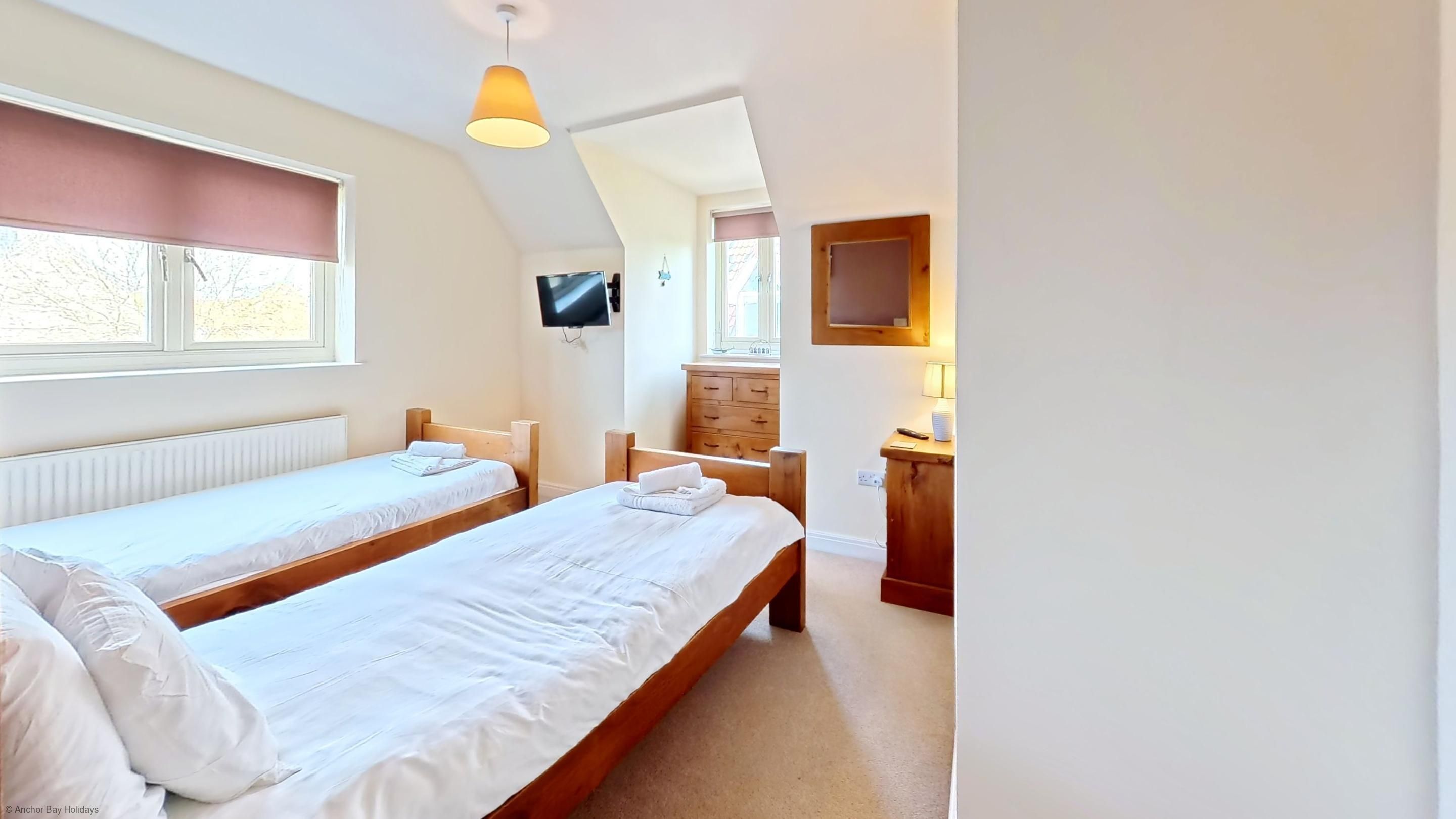 Une chambre avec deux lits simples, une commode en bois, un téléviseur fixé au mur et une fenêtre donnant sur l’extérieur.