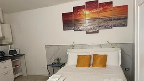 Quarto de hotel com cama, escrivaninha com computador e pintura de um pôr do sol sobre o oceano na parede.