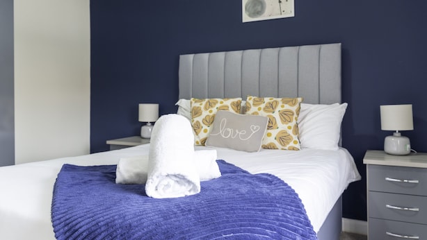 A neatly made bed with a rolled white towel, a purple blanket, and a pillow with a heart design. There are two bedside tables with lamps and a wall-mounted light fixture. - Raploch House (Larkhall)