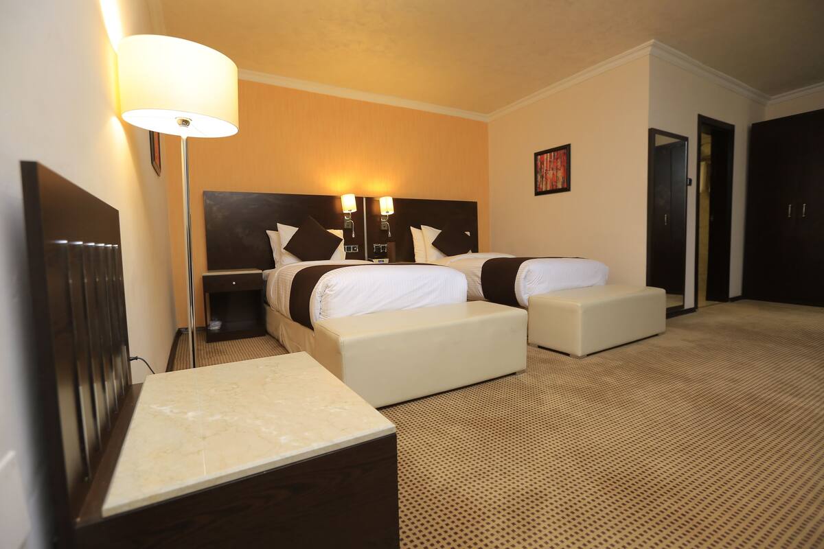 A hotel room with two beds, a desk, a lamp, and a painting on the wall.