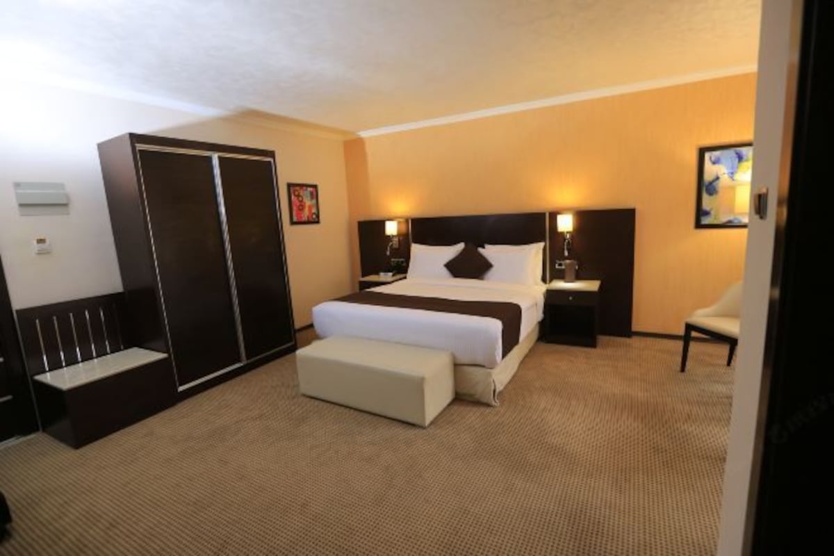 A hotel room with a large bed, a bedside table, a nightstand, a wardrobe, and a bench.