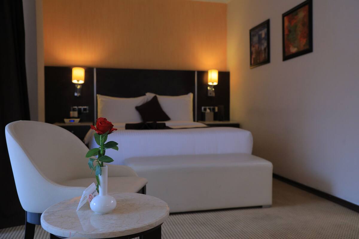 A hotel room with a bed, a white armchair, a small round table with a vase of a red rose, and two wall-mounted lamps.