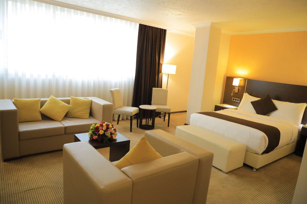 A hotel room with a bed, a sofa, a chair, a small table with a flower arrangement, and a lamp.