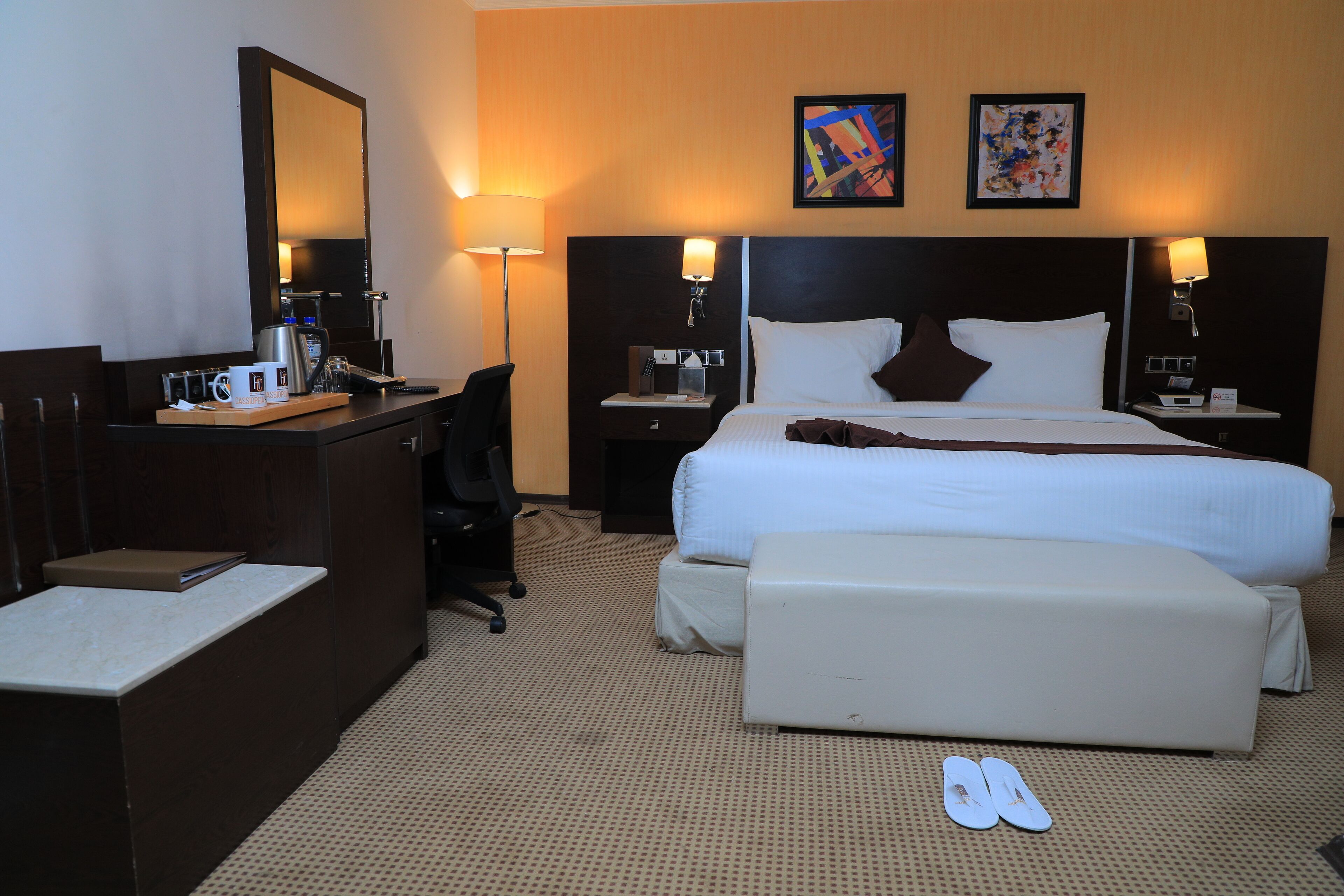 A hotel room with a large bed, a desk with a coffee maker, a chair, and a pair of slippers on the floor.