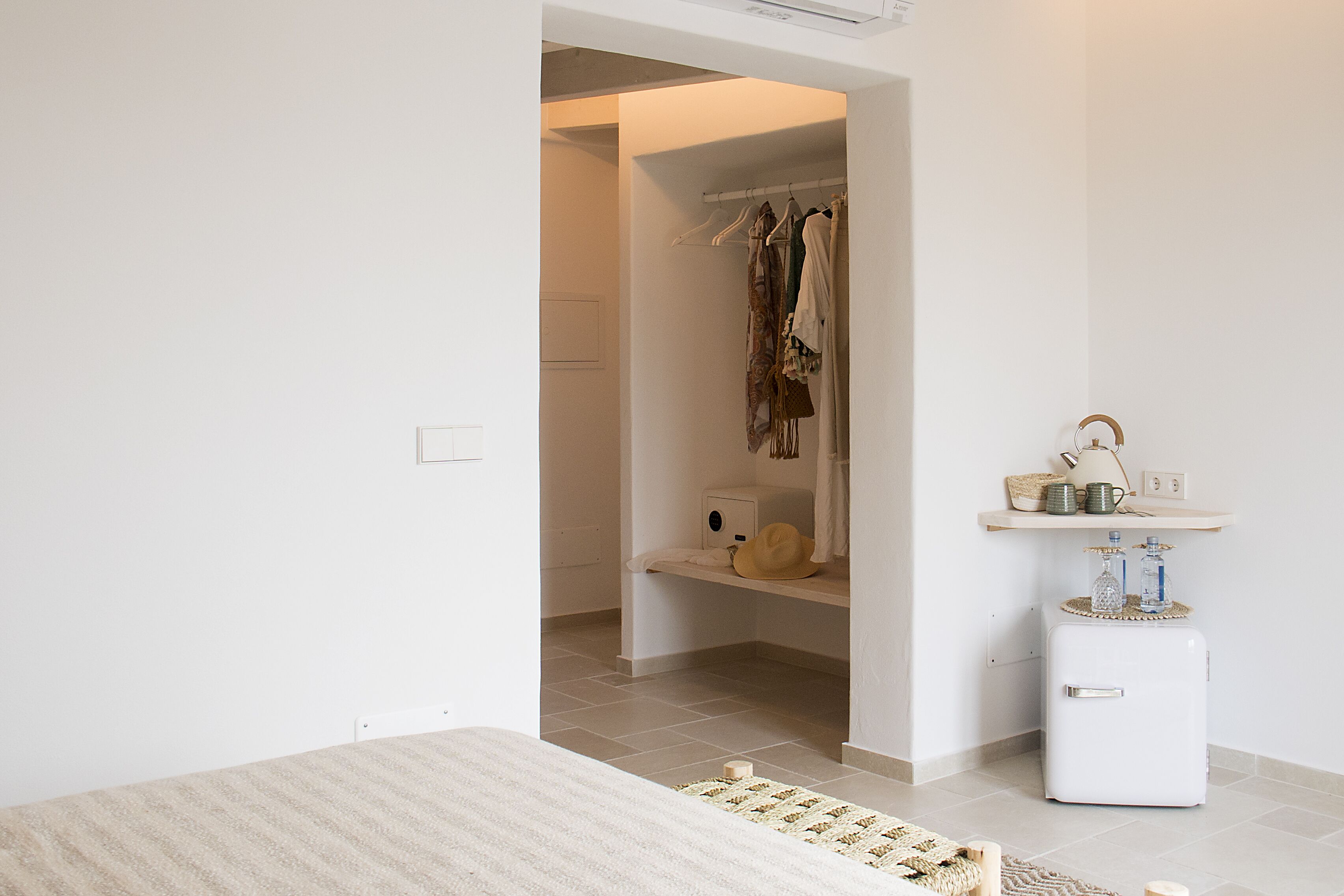 Une chambre à coucher avec un lit, une armoire où sont suspendus des vêtements, une petite commode blanche et une table de chevet avec une lampe et des objets décoratifs.