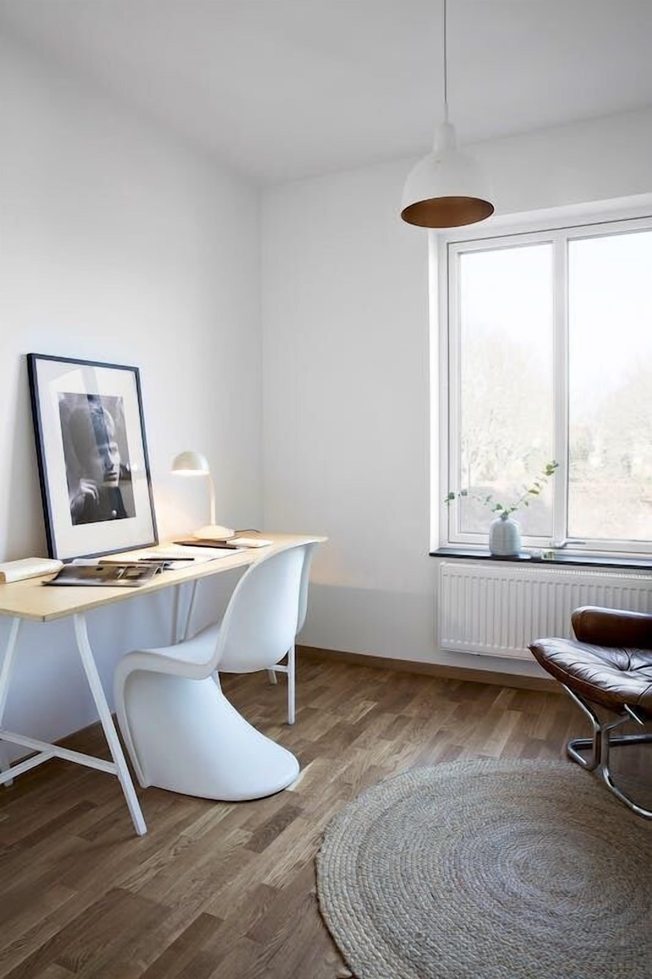 A modern, minimalist office space with a wooden desk, white chairs, a hanging pendant light, and a view of the outdoors through a large window.
