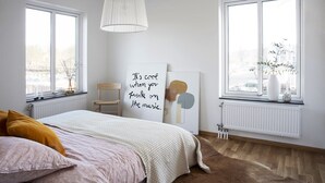 Un dormitorio con cama, dos ventanas, un radiador, una silla y una pared con un cuadro enmarcado.