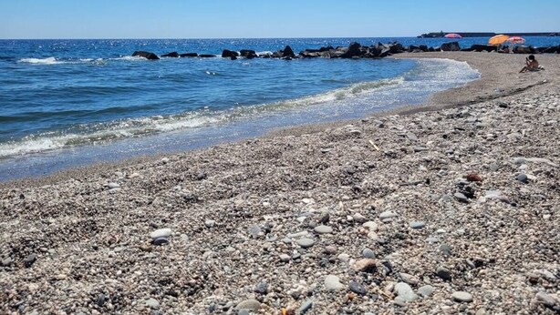 Una spiaggia di ciottoli con cielo azzurro limpido, mare calmo e alcuni ombrelloni in lontananza.