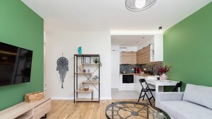 A modern living room with a sofa, a glass coffee table, and a TV on the wall. There is a kitchen area in the background with wooden cabinets and a dining table.