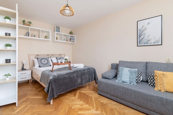 A modern bedroom with a bed, a grey sofa, wooden flooring, and built-in shelving with plants and books.