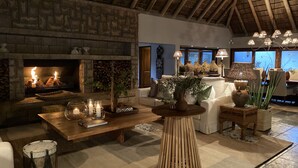 A cozy living room with a stone fireplace, wooden furniture, and a high ceiling with a thatched roof. - Simbavati River Sands (Bushbuckridge)