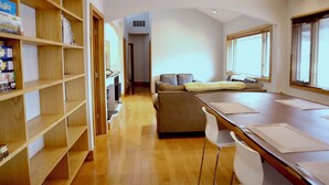 A modern, well-lit room with a wooden floor, a sofa, a dining table, and a bookshelf.