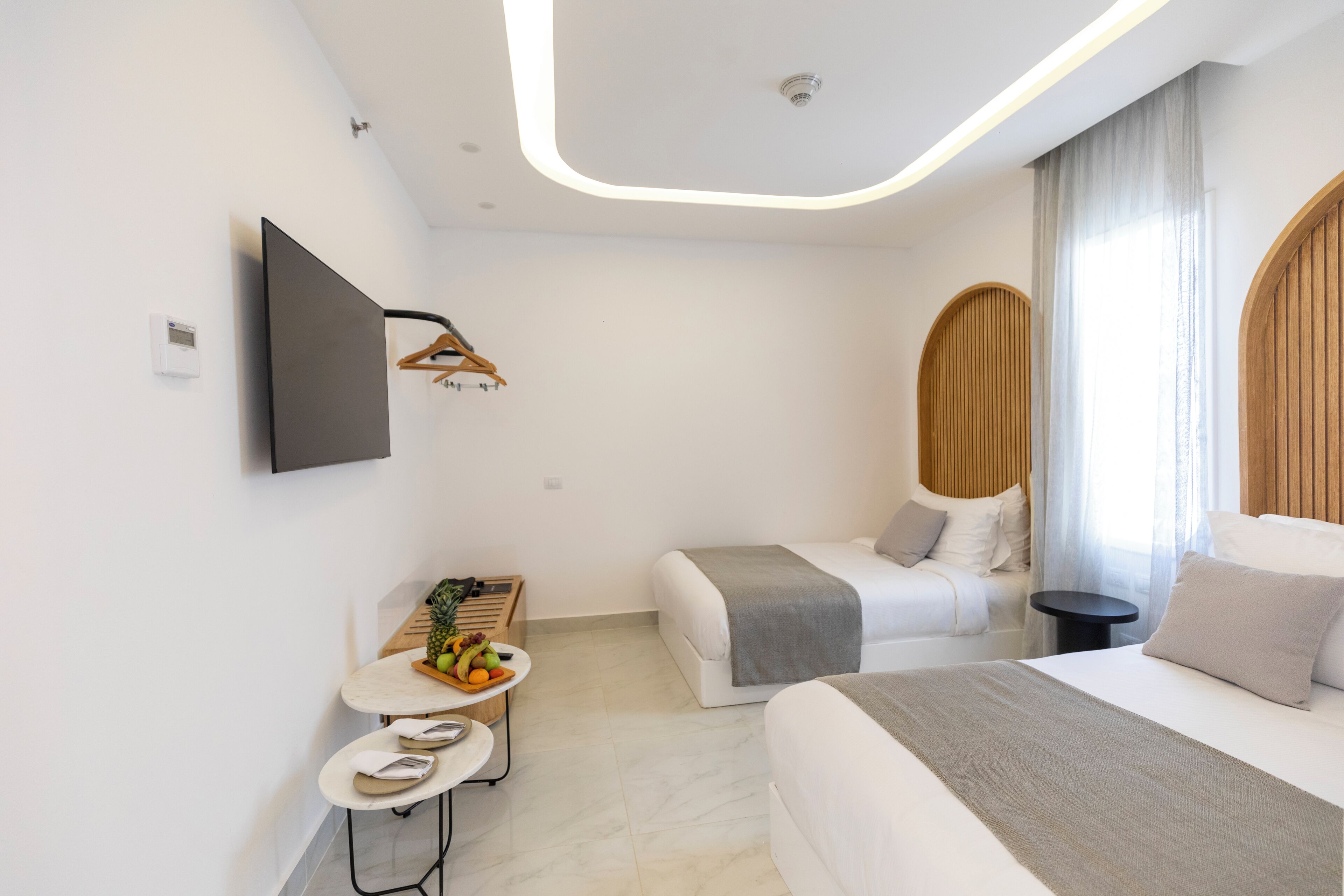 Quarto de hotel moderno com uma cama grande, uma cama menor, uma TV de tela plana na parede, uma mesinha redonda com uma cesta de frutas e uma mesinha lateral redonda com um par de chinelos.