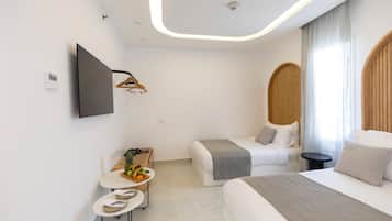 Quarto de hotel moderno com uma cama grande, uma cama menor, uma TV de tela plana na parede, uma mesinha redonda com uma cesta de frutas e uma mesinha lateral redonda com um par de chinelos.