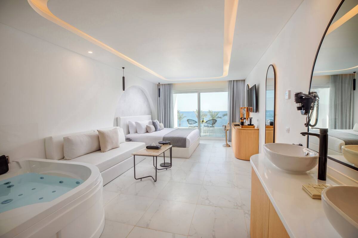 A modern hotel room with a large bathtub, a sofa, a dining table, and a large mirror.