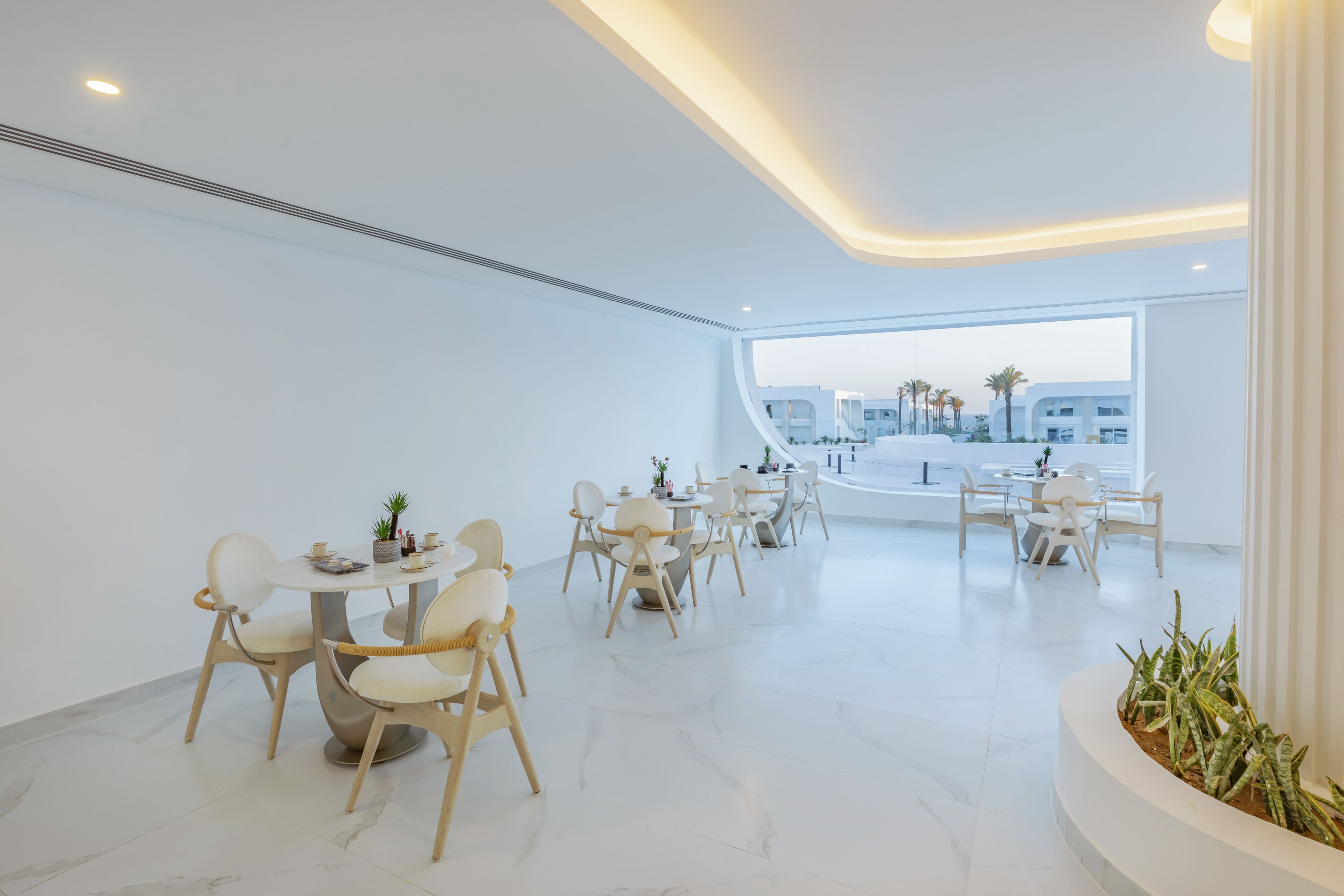 Un comedor moderno con mesas y sillas redondas, un ventanal con vistas a edificios y palmeras, y una planta en maceta a la derecha.